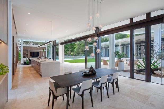 A spacious, light-filled open-plan dining and kitchen interior with floor-to-ceiling glazing overlooking a landscaped courtyard. The room features a long dark dining table with contemporary chairs, a large stone island, pale stone flooring, exposed dark timber or metal structural beams, and a sculptural cluster pendant light.