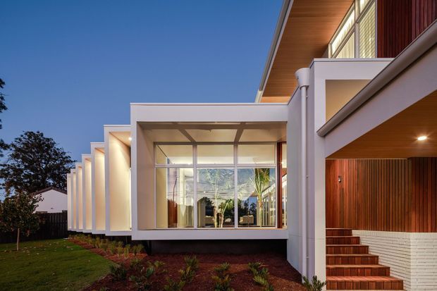 Exterior architectural photograph of a contemporary multi-level residence at dusk. The facade combines crisp white rendered volumes, extensive floor-to-ceiling glazing, warm timber soffits and vertical timber cladding, with a covered entry terrace and tiled steps. Landscaped planting and lawn sit alongside the building.