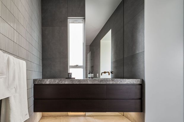 A contemporary bathroom viewed head-on, featuring a floating dark wood vanity with a broad grey stone benchtop, large dark grey wall tiles, a narrow vertical window, wall mirror, chrome tapware, and a light tiled floor. A towel rail with white towels is visible on the left.