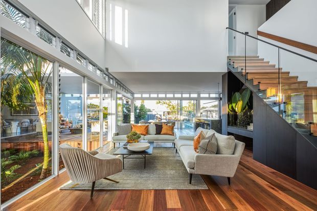 Bright contemporary double-height living room with extensive floor-to-ceiling glazing, a visible waterfront or marina outlook, warm timber flooring, neutral upholstered seating, and a sculptural open staircase with glass balustrades and dark built-in joinery. The space is airy and open, with indoor planting and strong indoor-outdoor connection.
