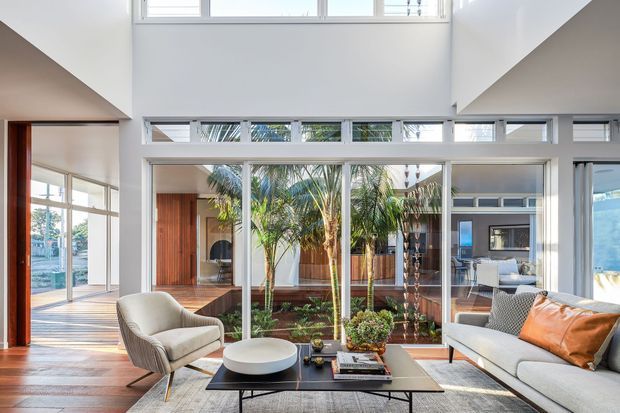 Bright contemporary open-plan living room with a double-height ceiling and extensive white-framed glazing overlooking a planted internal courtyard. The space features warm timber flooring and wall elements, a pale upholstered sofa and lounge chair, a black low coffee table, and restrained modern furnishings. A dining or secondary living area is visible beyond the glazing, with strong indoor–outdoor visual connection and abundant natural light.