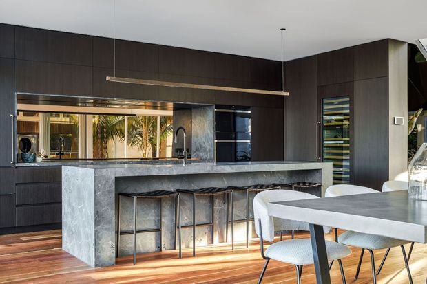 Contemporary open-plan kitchen and dining area with extensive dark timber cabinetry, a long grey stone island with integrated seating, stainless-steel appliances, and a large dining table. Warm timber flooring contrasts with the dark joinery, while a linear pendant light and indoor-outdoor glazing create a refined, modern atmosphere.
