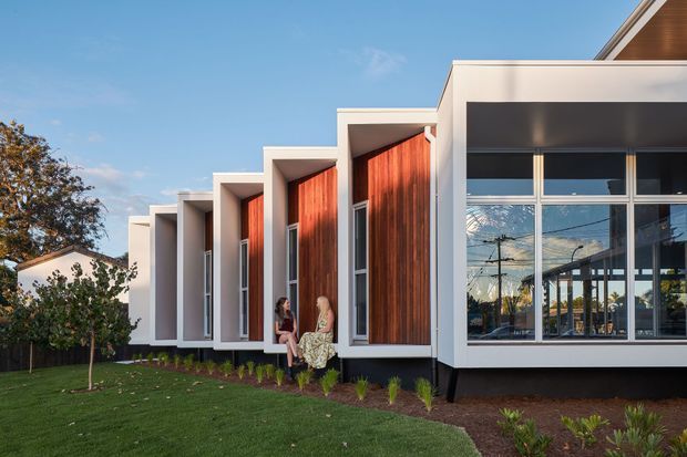 A contemporary low-rise building exterior with a rhythmic series of projecting white rectangular bays, warm vertical timber-clad recessed panels, and tall narrow windows. A larger glazed frontage occupies the right side, reflecting the surrounding suburban street. The building sits beside a neatly landscaped lawn with young planting and mature trees, under a clear blue sky.