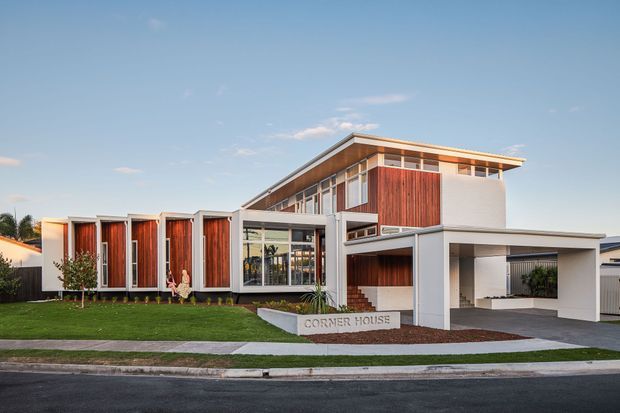 A modern multi-level residential house viewed from the street, with strong horizontal rooflines, projecting upper volume, extensive glazing, white rendered walls, and warm vertical timber-clad feature panels. The frontage includes a manicured lawn, concrete footpath, landscaped garden beds, and an integrated double carport/garage area. A sign identifies the property as “Corner House.”