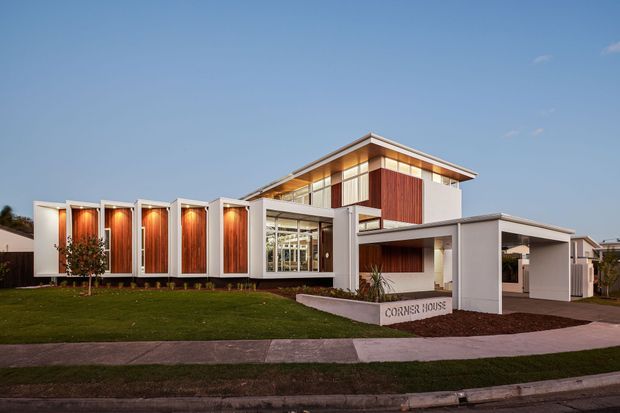 Exterior architectural photograph of a contemporary two-storey residence at dusk, with a strong linear composition of white rendered volumes, warm vertical timber-clad wall panels, extensive glazing, deep roof overhangs, and a covered carport. The home sits behind a lawn and footpath, with a low garden wall bearing the text “CORNER HOUSE”.