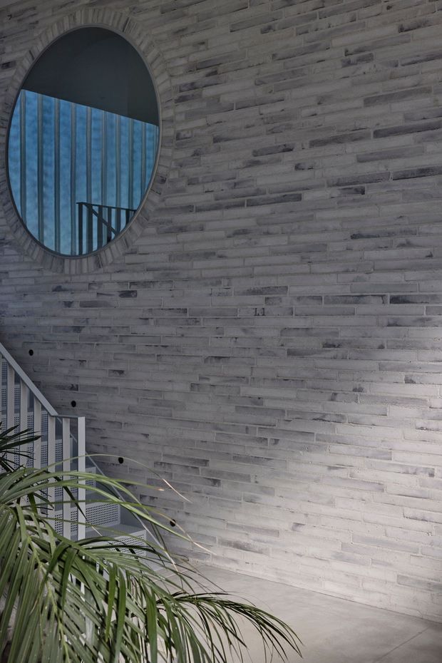 Interior architectural detail showing a curved light-grey brick wall with a large circular window, visible blue-toned glazing and dark stair railing beyond, plus a white metal stair and tropical planting in the foreground. The space has a cool, minimalist, contemporary character with strong geometric forms and textured masonry.