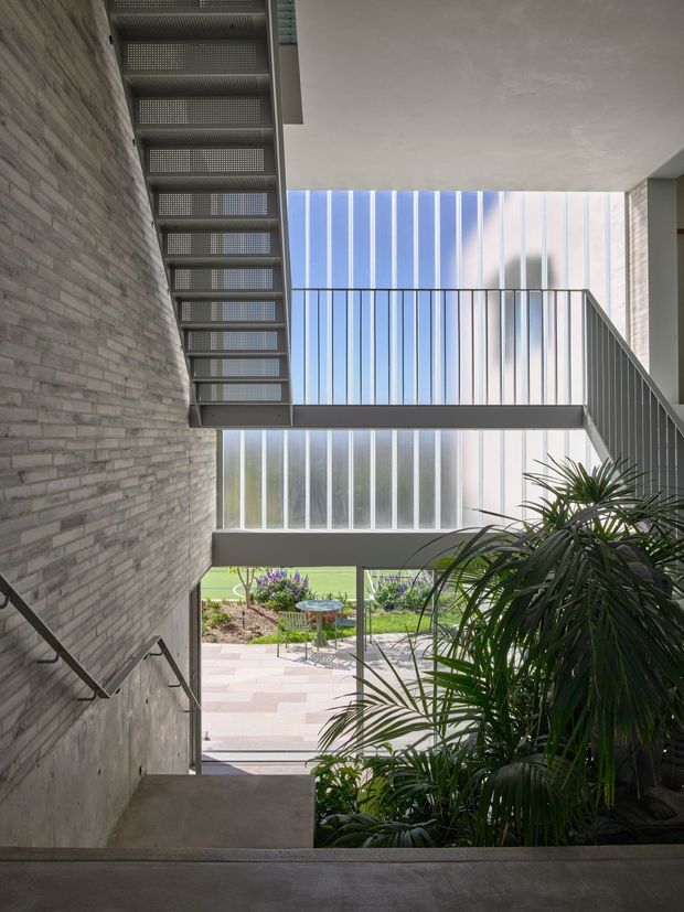 A contemporary multi-level stairway viewed from within a light-filled interior atrium. Exposed grey brick walls and concrete stair landings frame open metal stair flights and slender metal balustrades. Vertical translucent glazing fills the rear facade, bringing in blue daylight and views to a landscaped courtyard and paved outdoor area. Lush indoor planting occupies the lower right foreground.