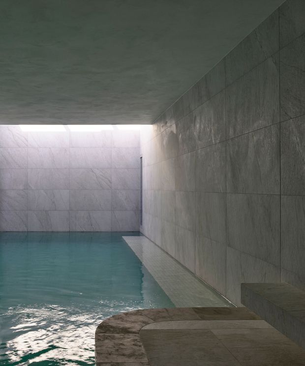 A moody, minimalist indoor swimming pool enclosed by large grey stone or porcelain wall tiles. The pool extends along the left side, with a narrow tiled edge and stepped built-in seating or ledge on the right. A low ceiling and linear opening at the far end admit soft natural light, creating a serene, spa-like atmosphere.