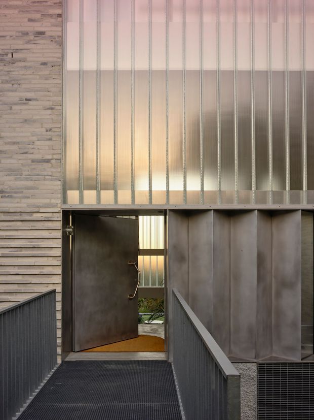 A close architectural view of a contemporary building entrance approached by a narrow metal-grating ramp or bridge. The facade combines horizontally layered stone cladding on the left with large vertical translucent and reflective metal or glass panels above and beside a substantial dark metal door. Slim metal railings, an industrial floor grate, and a partially visible planted courtyard beyond the open doorway reinforce the modern industrial character.
