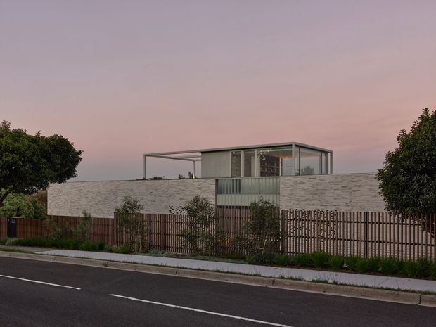 Exterior view of a low-profile contemporary residence from across a suburban street. The home has a long pale masonry or stone-textured wall, a rooftop box-like pavilion with extensive glazing and slender dark metal frames, and timber vertical-slat fencing with landscaped planting in front. The flat roofline and open upper-level terrace create a minimalist modern composition, photographed at dusk under a pink sky.