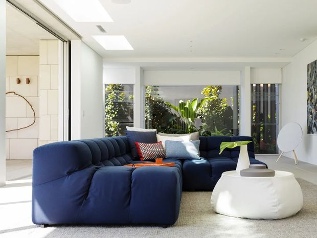 Bright contemporary living room with a large deep navy-blue sectional sofa, white sculptural coffee table, neutral carpet, and minimalist white walls. Full-height glazing and skylights bring in abundant daylight, with leafy greenery visible outside. Built-in pale cabinetry and an abstract artwork add subtle visual interest.