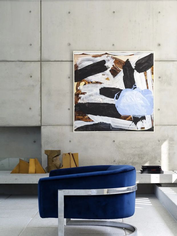 A contemporary interior vignette featuring a deep blue curved velvet lounge chair with a polished metal frame, positioned against a gridded exposed concrete wall. An abstract black, white, brown, and pale blue artwork hangs above a low concrete bench with sculptural timber objects. Strong sunlight creates striped shadows across the pale floor and steps.