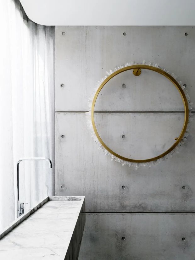 A minimalist bathroom vanity area with a white marble countertop and integrated basin at left, a slim chrome wall-mounted tap, sheer curtains, and a large round mirror with a brass-gold frame and textured clear decorative edge. The backdrop is exposed grey concrete wall panels with visible fasteners and horizontal joints. The space is bright, restrained, and industrial-luxe in character.