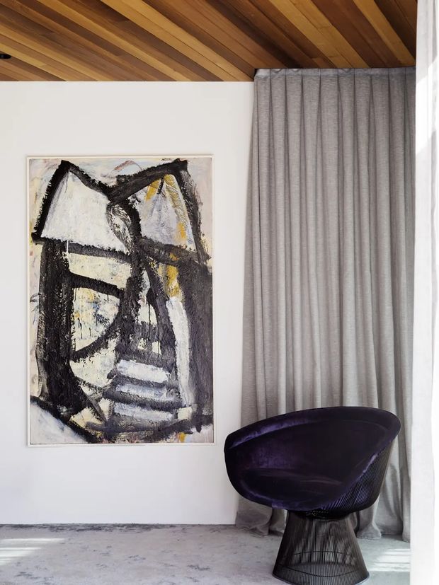 A refined interior corner with a dramatic abstract painting on a white wall, a sculptural deep-purple lounge chair, full-height grey curtains, and a warm timber-lined ceiling. The space has a restrained, gallery-like composition with strong material and colour contrast.