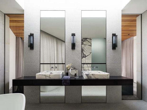 A refined contemporary bathroom vanity wall with two white vessel basins set on a long dark floating counter. Full-height mirrors, grey textured wall panels, black wall sconces, pale stone surfaces, and timber-lined recessed areas with curtains create a symmetrical, luxurious composition. A freestanding bathtub is partially visible in the foreground.
