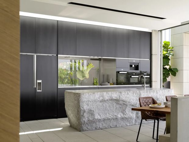 Contemporary open-plan kitchen with a dramatic dark full-height cabinetry wall, integrated appliances, reflective splashback, and a large sculptural pale stone island with a waterfall-style edge. A dining table and chairs occupy the foreground, while abundant natural light and indoor planting create a refined, high-contrast interior.