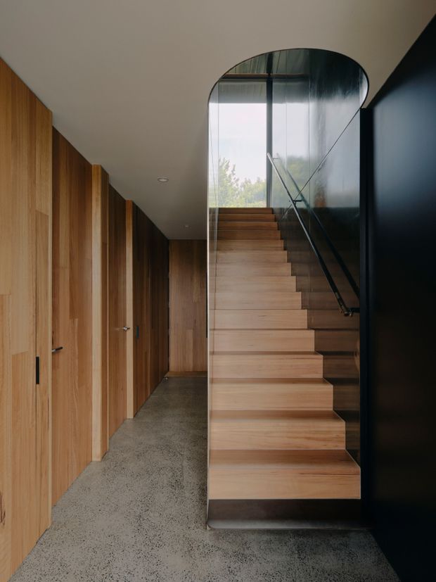 Interior view of a refined hallway and stairway. A straight timber staircase rises beside a clear glass balustrade, with a curved glazed enclosure at the upper landing. Full-height warm timber doors and wall panelling line the left side, while a dark painted wall frames the right. The floor is polished grey concrete or terrazzo, creating a restrained, modern material palette.