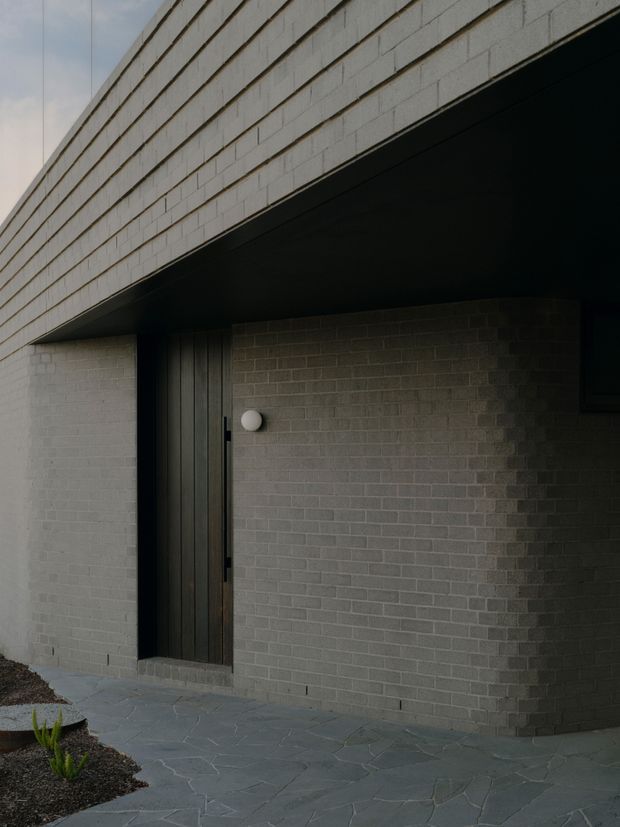 A minimalist modern building entrance beneath a deep black projecting roof canopy. The facade is constructed from pale grey masonry blocks, with a tall dark timber or metal vertical-plank door recessed into the wall. Irregular grey stone paving leads to the entry, bordered by a sparse landscaped garden. The composition emphasizes strong horizontal roof lines, a restrained monochrome palette, and dramatic shadow.