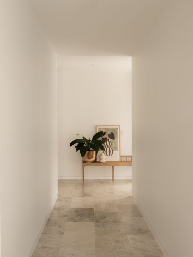 A narrow, softly lit minimalist hallway with warm off-white walls and a pale natural stone tile floor. At the end of the corridor is a simple timber console table styled with a leafy potted plant, framed artwork, ceramic objects, and a small framed print. The space is calm, uncluttered, and neutral in tone.
