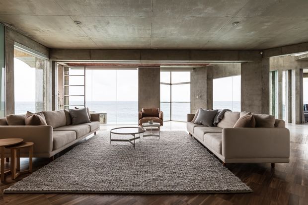 A spacious contemporary living room with two large neutral sofas facing each other across a textured area rug and low round coffee table. A leather lounge chair sits between expansive floor-to-ceiling glazing overlooking the ocean. Exposed concrete walls, columns and ceiling contrast with dark herringbone timber flooring, while the open plan and generous glazing create a refined, coastal atmosphere.