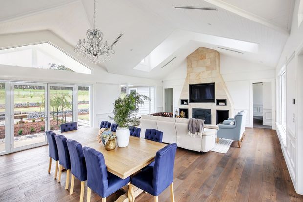 Bright open-plan living and dining area with white walls, vaulted ceilings, skylights, extensive glazing, dark timber flooring, and a stone-clad fireplace wall. A large timber dining table with blue upholstered chairs occupies the foreground, while a white sofa and armchairs create a seating area around the fireplace.