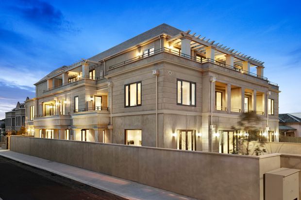Exterior twilight view of a large multi-storey residence with a symmetrical, classical facade. The building has pale rendered masonry, horizontal banding, columns, pilasters, balconies, extensive glazing, a pitched tiled roof, and rooftop pergola structures. Warm interior and facade lighting contrasts with the blue evening sky, while a high perimeter wall encloses the property.