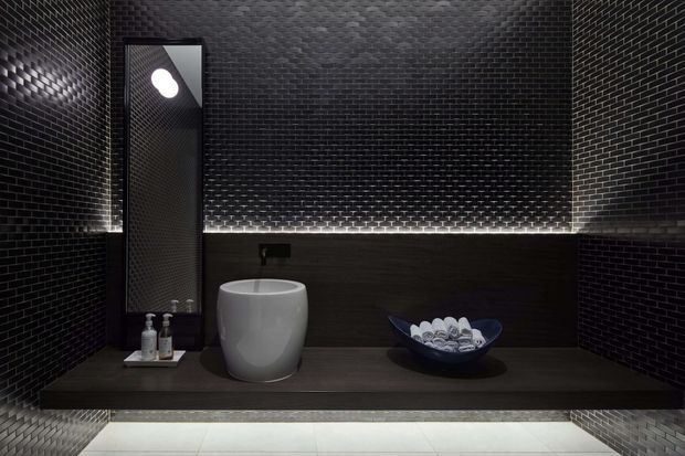 A dark, luxurious bathroom vanity with black textured mosaic wall tiles, a wall-mounted black basin tap, a white vessel basin, and a dark floating vanity shelf. A tall narrow mirror, toiletries, and a decorative bowl of rolled towels complete the composition. Subtle linear lighting illuminates the rear ledge and the pale tiled floor creates contrast.