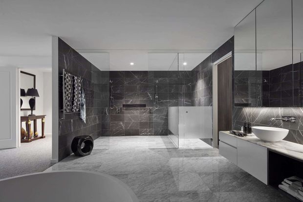 A spacious contemporary bathroom in a dark, monochromatic palette. Large-format charcoal stone-look wall tiles contrast with pale textured floor tiles and white minimalist cabinetry. The room includes a freestanding bathtub in the foreground, a wall-mounted vanity with a vessel basin, a large mirror, and a walk-in shower with clear glazing. An adjoining carpeted bedroom or dressing area is visible through the open doorway.