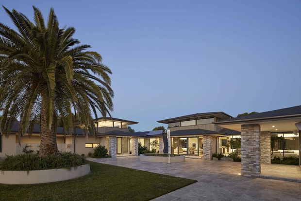 A twilight exterior view of a low-rise contemporary residence or complex arranged around a landscaped courtyard. The building features broad roof overhangs, extensive glazing, stacked-stone clad columns, pale rendered walls, and a large paved forecourt. A mature palm tree and lawn dominate the foreground.