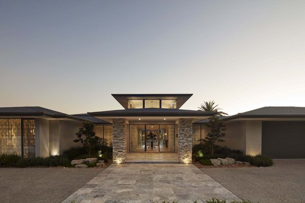 Symmetrical single-storey contemporary residence photographed at dusk, with a central entry pavilion, broad low-pitched rooflines, clerestory-like glazed upper volume, stone-clad entry walls, expansive glazing, landscaped beds, and a paved approach. The composition emphasizes the home's balanced facade and warm architectural lighting.