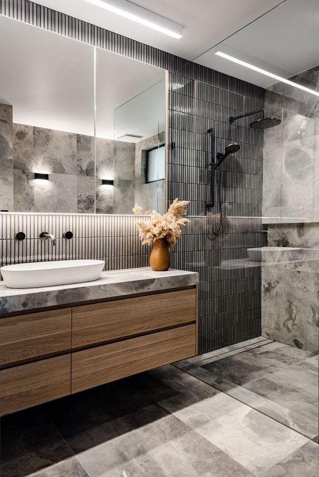 Contemporary bathroom featuring a floating timber vanity with a white vessel basin, large-format grey stone-look wall and floor tiles, a black vertically ribbed feature tile wall, matte black shower fittings, a frameless mirror, and linear recessed ceiling lighting. Neutral grey, white, and warm timber finishes create a refined, modern look.