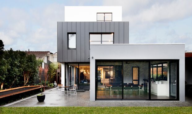 Exterior view of a contemporary multi-level urban residence with a strong geometric composition. The home combines dark vertically ribbed cladding, smooth white rendered walls, extensive black-framed glazing, and a flat roofline. A glazed ground-floor living area opens toward a landscaped yard with a raised timber walkway and outdoor seating. A neighbouring pitched-roof house is partially visible behind the property.