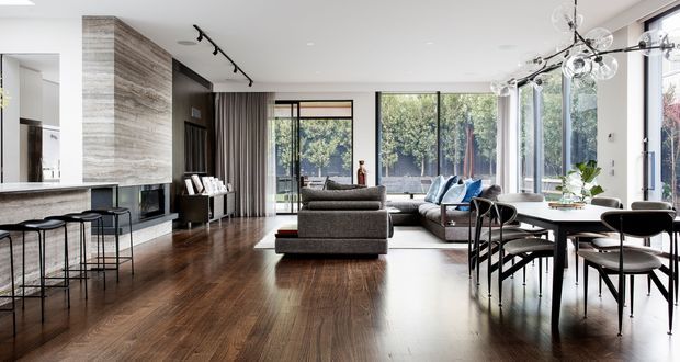 Open-plan contemporary living and dining area with a kitchen island at left, dark timber flooring, a large sectional seating arrangement, and a dining table beside extensive floor-to-ceiling glazing. The room has a bright, refined feel with dark built-in joinery, neutral furnishings, and sculptural modern lighting.