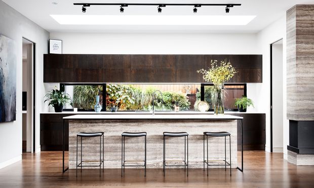 A bright contemporary kitchen centered on a long island with four slim black bar stools. The room features dark timber full-height cabinetry, a pale stone-look island benchtop and waterfall end, integrated shelving and a long horizontal window overlooking greenery. White walls and ceiling, warm timber flooring, a large skylight, and black track lighting create a clean, refined space.