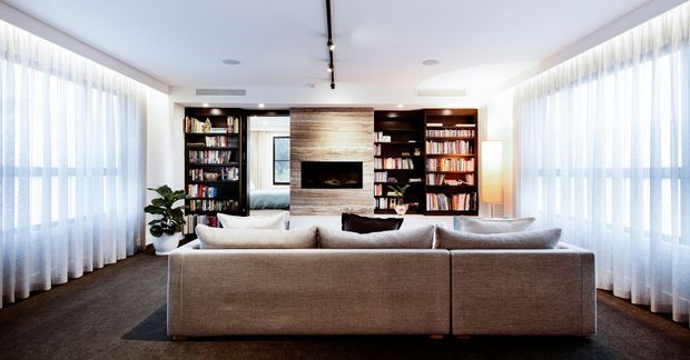 Bright contemporary living room dominated by a large modular sectional sofa, with full-height sheer curtains across expansive windows, dark built-in bookcases, and a central stone or timber-textured fireplace wall with open shelving. The room has a clean, refined fit-out and balanced neutral palette.