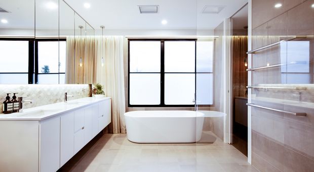 Bright contemporary bathroom with a freestanding white bathtub centered beneath large black-framed windows, a long white double vanity on the left, and a glass-enclosed shower with pale stone wall finishes on the right. Mirrored wall panels reflect the vanity and windows, while sheer curtains and warm timber detailing add softness.