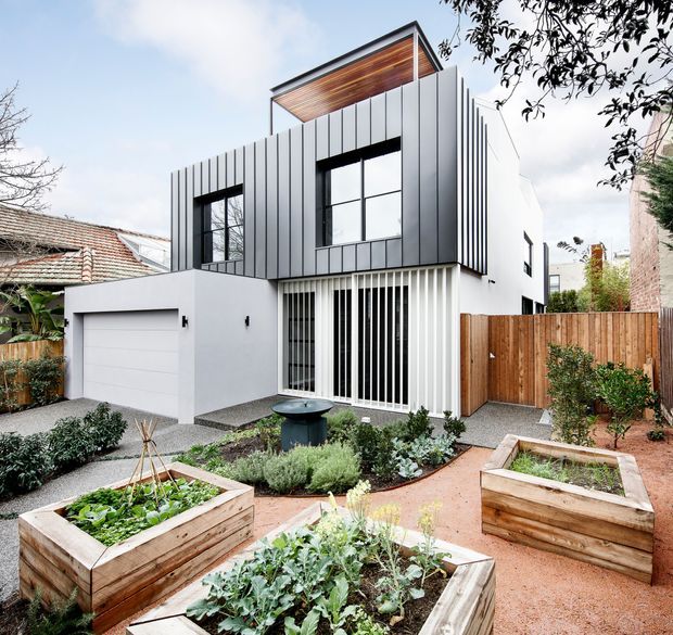 Contemporary multi-level urban residence viewed from the landscaped front garden. The house has a crisp white rendered lower level, dark grey vertical metal-clad upper volume, expansive black-framed windows, a projecting roof terrace or canopy lined with timber, and a white garage. Raised timber garden beds, gravel paths, timber fencing, and established planting occupy the foreground.