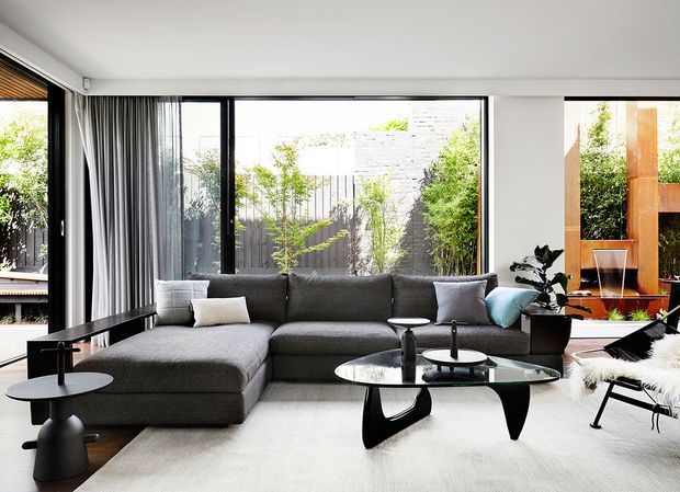 A refined contemporary living room with a large dark grey L-shaped sectional sofa, black sculptural coffee tables, and a pale area rug. Full-height black-framed glazing opens views to a planted courtyard, while floor-to-ceiling grey curtains, a potted plant, and a modern lounge chair complete the calm, monochromatic interior.