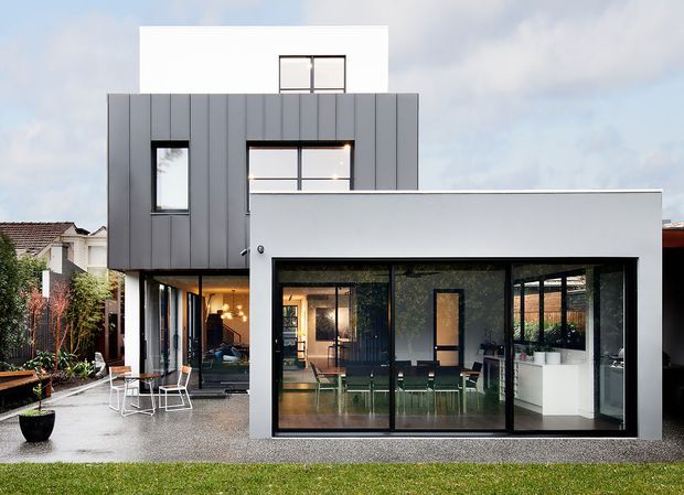 Exterior view of a contemporary multi-level residential extension or townhouse, featuring a rectilinear white-rendered volume, dark grey vertical metal-like cladding, and extensive black-framed glazing. The ground floor opens onto a paved courtyard with outdoor seating, while interior dining and kitchen areas are visible through the glass. A neighbouring traditional tiled-roof house is partially visible at left.
