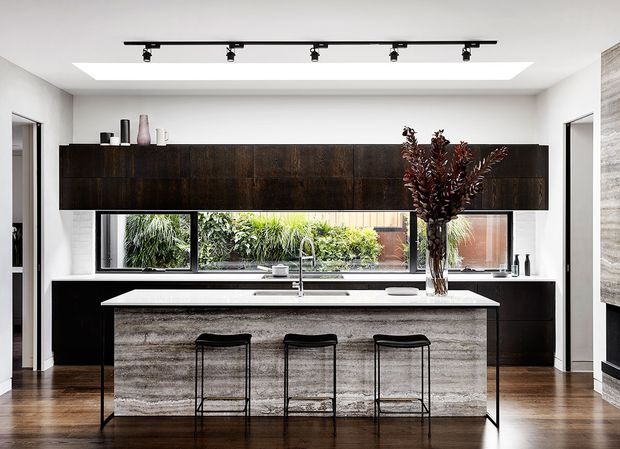 A contemporary kitchen dominated by a large central island with three bar stools. Dark timber cabinetry spans the rear wall above a long benchtop and integrated sink, while a textured grey island front contrasts with the dark cabinetry and dark timber floor. A horizontal garden-facing window runs behind the sink, and a large skylight with black track lighting brightens the room.