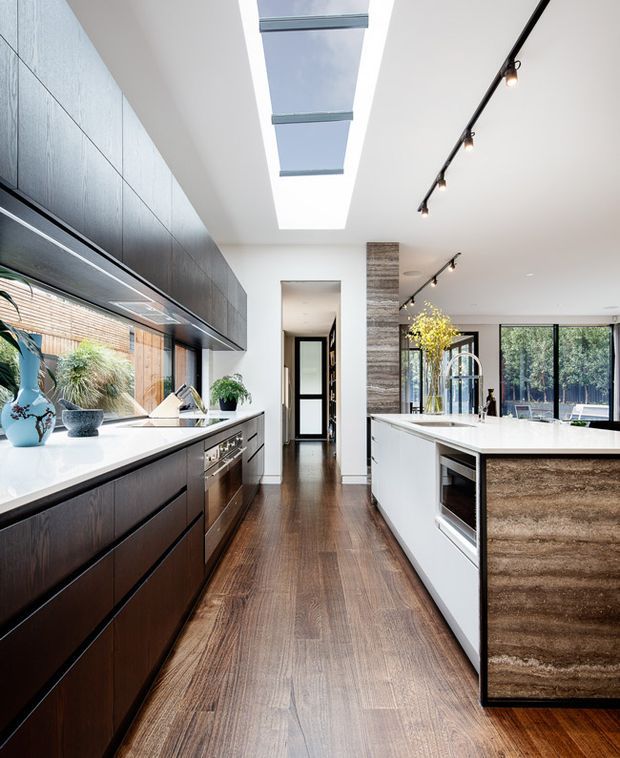 A bright, contemporary galley kitchen with a long dark timber island and matching full-height cabinetry, white benchtops, integrated appliances, and a timber flooring. A large linear skylight brings daylight into the space, while black track lighting provides additional illumination. Glazed doors and windows open the kitchen toward a garden, and a central passage leads to the rest of the home.