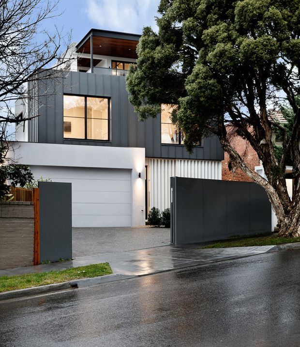 Street-facing view of a contemporary multi-storey residence with dark vertical metal cladding, large black-framed windows, a white rendered lower level, integrated garage, tall privacy gates, and a sheltered upper terrace with a timber-lined soffit. Mature trees partially screen the facade, and the wet pavement creates reflective highlights.