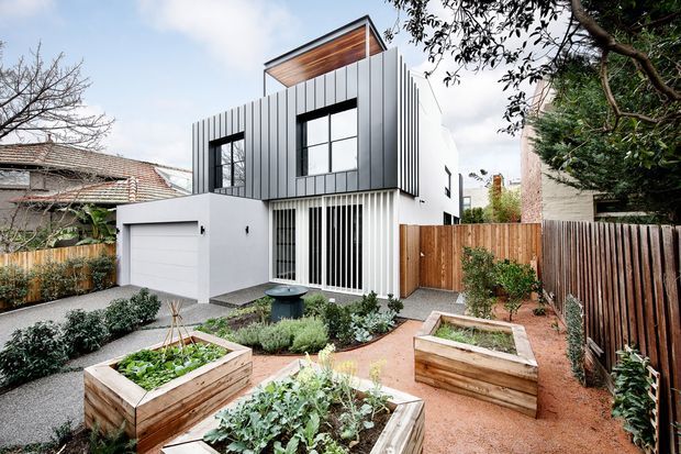 Contemporary two-storey urban residence viewed from a landscaped front garden. The facade combines crisp white rendered walls with dark vertical metal cladding, large black-framed windows, a projecting timber-lined roof feature, and a white integrated garage. Raised timber garden beds, gravel paths, low planting, and timber boundary fencing define the foreground courtyard.