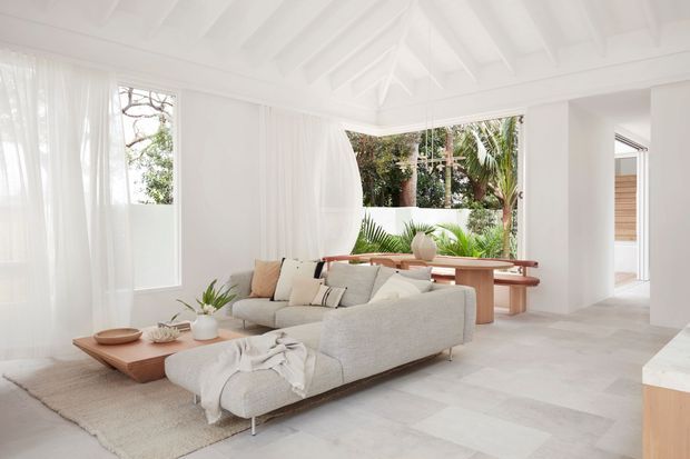 Bright, airy open-plan living room with white walls, sheer curtains, exposed painted ceiling beams, pale stone flooring, and a large light neutral sectional sofa. A timber coffee table and built-in timber dining bench add warmth, while wide openings connect the room to lush tropical greenery and an adjacent space.