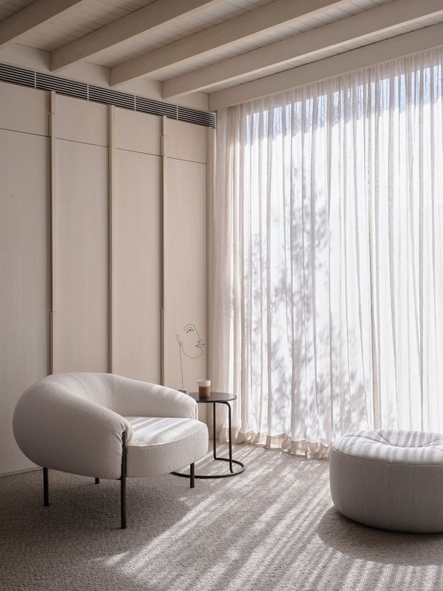 A softly lit minimalist sitting area with a sculptural boucle armchair, matching rounded ottoman, slim black side table, and pale full-height sheer curtains. The room features pale vertical built-in wall panelling, exposed painted ceiling beams, integrated linear ventilation, and a neutral textured carpet. Strong daylight filters through the curtains, creating striped shadows across the floor.