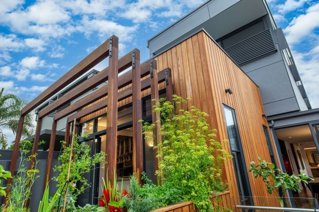 Low-angle exterior view of a contemporary multi-level residence with warm vertical timber cladding, dark charcoal metal elements, and a prominent timber pergola or screened entry structure. Large glazed openings reveal part of the interior, while layered planting and a raised garden edge soften the facade. The image is brightly lit beneath a partly cloudy blue sky.