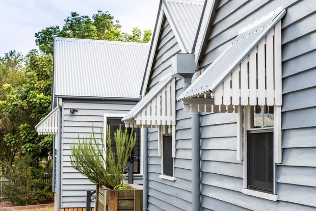 Exterior view of a grey weatherboard-clad home with steep corrugated metal gable roofs, white-trimmed windows, and distinctive white slatted window awnings. A timber planter and garden vegetation sit beside the facade.