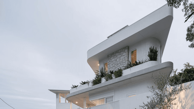 Exterior view of a sculptural multi-level residential building with sweeping curved slab edges, white ribbed cladding, pale stone masonry, warm timber-toned window accents, and planted balconies. The overcast setting and surrounding vegetation frame the modern facade.