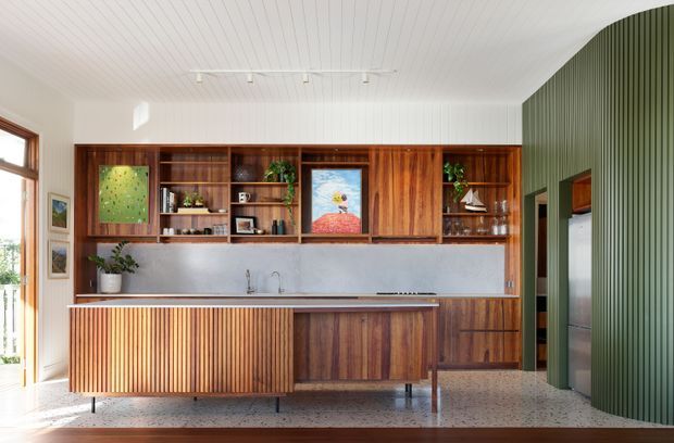 Bright, carefully detailed kitchen featuring a long timber island with vertical fluted paneling, extensive dark timber cabinetry and open shelving, a pale stone backsplash and benchtop, and a terrazzo-style floor. The room has a white linear-panel ceiling, a strongly curved green ribbed feature wall, warm timber-framed glazing, and integrated display shelving with plants and artwork.