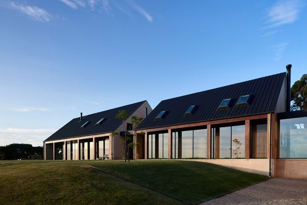 Exterior view of a contemporary, low-profile residential building with connected gabled roof volumes, dark standing-seam metal roofing, extensive floor-to-ceiling glazing, and warm timber-framed facade elements. The building sits in a broad lawned setting under a clear blue sky.